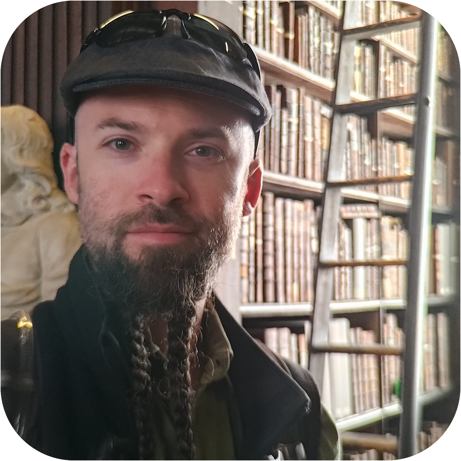 Josh Haas stares directly into the camera, slightly on the left side of the image. Behind him and on the right is a tall and wide bookshelf full of books, and a ladder going to the top.